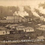 Sepia-toned vintage photo of a large Eatonville, WA sawmill or industrial timber complex with smoke billowing from chimneys, surrounded by early residential houses.