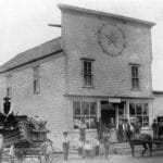 Black and white photo of a historic wooden general store building in early Auburn, Washington, with a large clock face; men in work clothes and horses with carriages stand outside, reflecting the town's early rail and agriculture workforce.